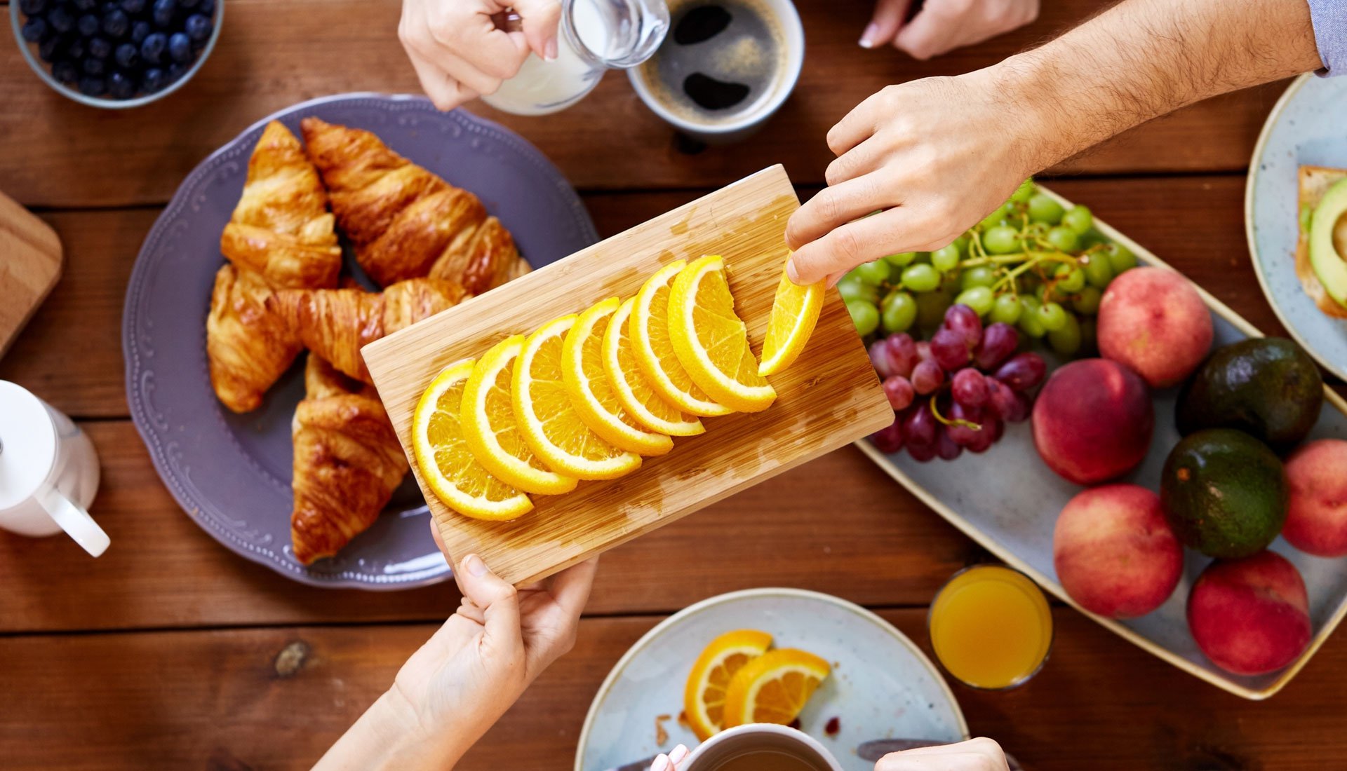 Copioso desayuno sobre una mesa de madera con una bandeja con rodajas de naranja en primer plano siendo degustada por una persona.